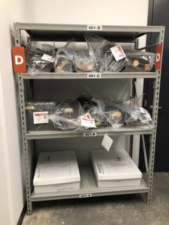 Grey metal shelving unit marked “D” which has radioactive artifacts stored on it. Artifacts are stored in white boxes on lower shelves, and are enclosed in clear plastic sheeting on the two upper shelves.