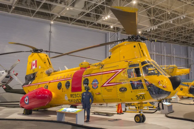 A huge yellow military helicopter sits on the floor of a museum. An information panel and a mannequin stand in front of it.