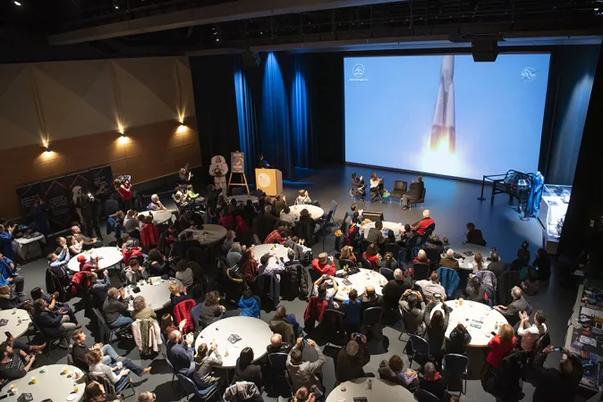 Plusieurs grandes tables rondes avec nappes blanches sont disposées dans une grande salle, et des gens y sont assis. À l’avant de la salle, trois personnes sont assises sur de grands fauteuils et parlent en pointant du doigt un écran sur lequel on peut voir une image d’un lancement de fusée.