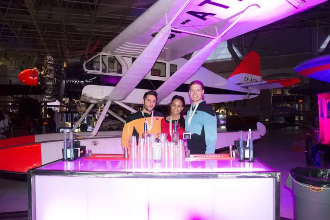 Three people wearing matching lanyards and Star Trek costumes are standing behind a table filled with clear plastic cups and a couple of drink bottles. Behind them sits a small seaplane and the room is lit with magenta lights.