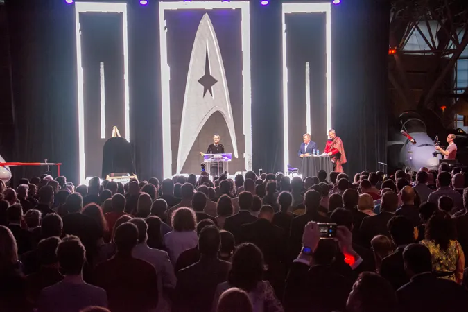 Une grande foule fait face à une scène avec des rideaux noirs. Trois personnes sont debout sur la scène et s’adressent au public. Derrière elles se trouvent des petites lumières argentées qui forment différents motifs, dont le symbole de Star Trek. 