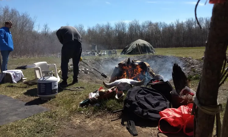 A person tends to an outdoor campfire fire, with a view of one of the Tipis and Telescopes sweatlodges in the background.