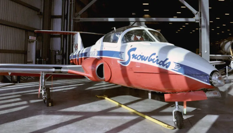 A white, blue, and red jet airplane, with the word “snowbirds” painted in blue along the front, sits on the floor of a warehouse. 