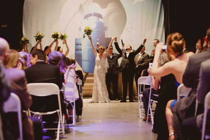 The scene of a wedding in the train room at the Canada Science and Technology Museum. The bride and groom have their hands up in celebration at the front of the room while multiple attendees watch from white fold-out chairs in rows across the room. Above the officiant is a large white cloth featuring the faint outlines of a steamboat.