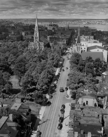 Une photo en noir et blanc prise du haut d’une rue de la ville montrant les rails de tramways, un tramway et plusieurs voitures. En arrière-plan, on voit une grande église et le havre d’Halifax.