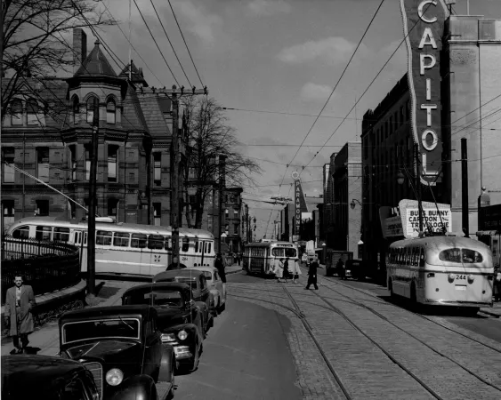 A black and white photograph of two streetcars on a city street, with people and 1940s-style cars parked nearby.