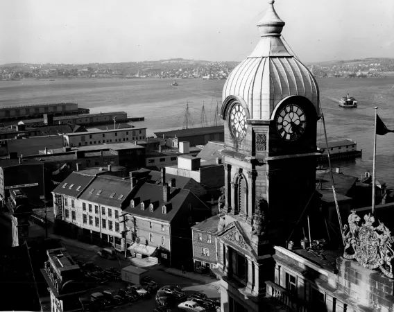 : A black and white photograph taken from the top of a building. In the foreground is a tall, dome-roofed tower, two flags, and the roof of the Dominion Customs building. In the mid-ground there are several smaller warehouse structures. In the background is Halifax Harbour.
