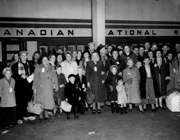 A black and white photograph of a large group of men, women and children. Many are holding bags and suitcases. They stand in front of a sign that reads “Canadian National Railway”.