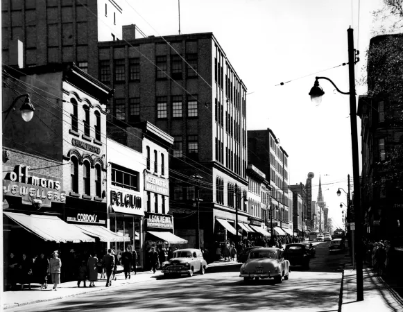 : A black and white photograph of a city street, with pedestrians walking on the sidewalk. Businesses line the street. There are several 1950s-style cars on the road.