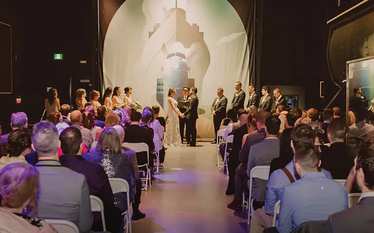 The scene of a wedding in between two trains at the Canada Science and Technology Museum. The bride and groom are holding hands at the front of the room while multiple attendees watch from white fold-out chairs in rows across the room. The bridesmaids and groomsmen are lined up at the front of the room. Above the officiant is a large white cloth featuring the faint outlines of a steamboat.