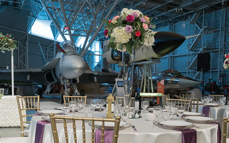 Part of the room at the Canadian Aviation and Space Museum which is large enough to fit a couple small planes in a corner. Multiple round tables with gold-backed chairs are scattered around the room. The tables are draped in lush white tablecloths and are decorated with bright pink, purple, and white flowers. There are big windows letting in light high up near the ceiling in the back of the room. 
