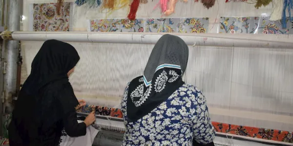 A mother and daughter weaving a carpet from a plan located on top of the loom