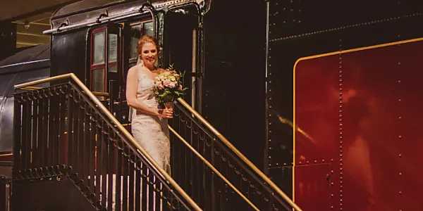 Une mariée tenant un bouquet de fleurs descend d’un des trains du Musée des sciences et de la technologie du Canada en empruntant un escalier avec garde-fous noirs et main courante dorée. Un homme en habit l’attend en bas de l’escalier. Le début du mot « Canadian » est visible sur le côté du train.