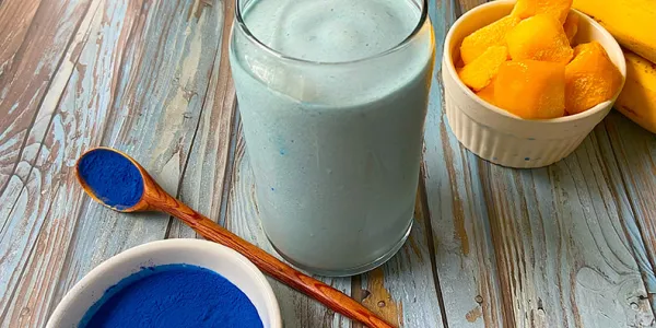 A glass filled with a thick, blue beverage sits on a rustic, wooden surface. A bowl and spoon with blue powder sits in the foreground, with mangoes and bananas in the background.
