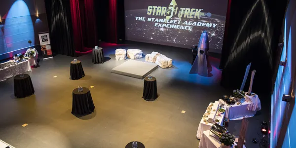Top down view of a room decorated with various space-themed items, small circular black tables scattered around the room and three long tables with white tablecloths filled with finger food on one side of the table. On the projector screen, the words “Star Trek the startfleet academy experience” is displayed.