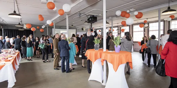 Groups of people standing around a large room with white ceiling and light walls and floors. There are small high tables scattered around the room decorated with orange tablecloths, flowers, and balloons