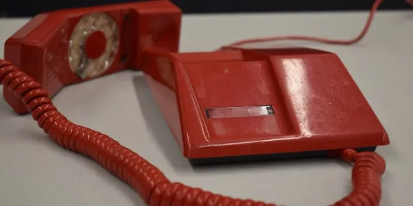 A red plastic telephone with the handset off of the base on a light grey table. There are scratches on the phone which is an angular design. The rotary dial is on the handset and attached to the base by a red spiral cord.