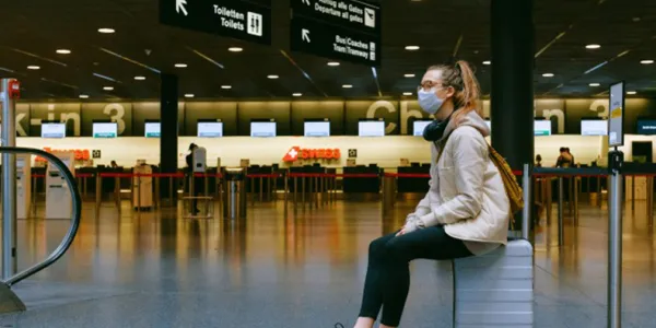 Inside an empty airport terminal, a young woman sits on her luggage while wearing a medical mask; she has headphones draped around her neck