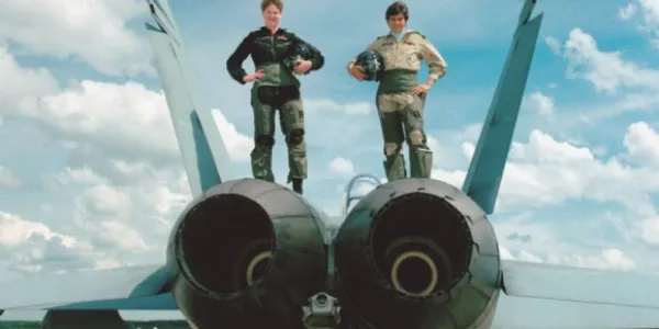 Deux femmes militaires en combinaison de vol posent avec confiance au sommet d'un avion de chasse sur une piste d'atterrissage par une journée claire et bleue.