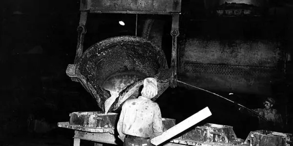 Image historique en noir et blanc montrant une énorme machine en bois dotée d’une immense cuve qui est basculée pour laisser couler du métal liquide dans des moules. Un homme, de dos, surveille les opérations.