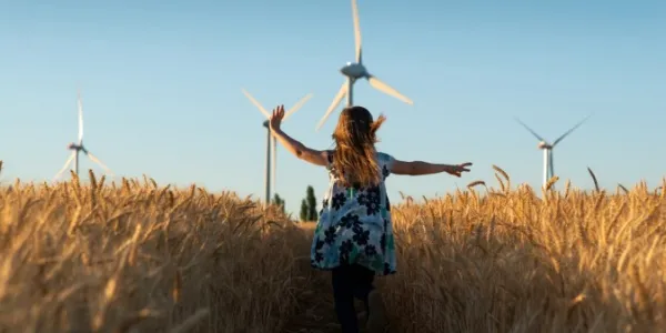 A young girl wearing a flowered dress is running through a path in a wheat field. Wind turbines are visible in the background, against a bright blue sky.