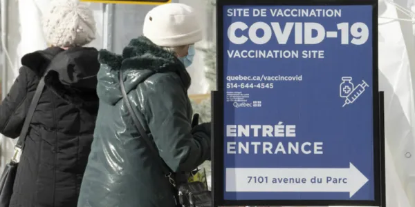 Two women in long winter jackets with white toques stand next to a COVID-19 vaccination site sign.