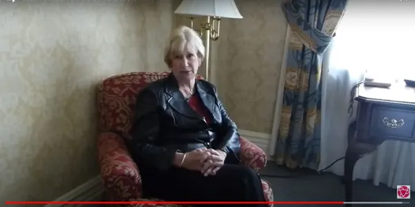 Arrêt sur image d’une vidéo; on voit une femme assise sur un fauteuil rouge, dans une pièce baignée de lumière naturelle.