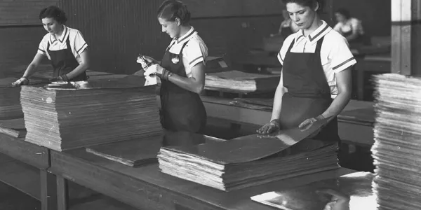 A black-and-white historical image shows three young girls wearing aprons over white shirts, standing in a row in front of a long, wooden desk. Several tall, square stacks of sheet metal stand in front of them.