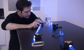 A person with dark hair and wearing a dark shirt uses equipment such as wooden blocks and mirrors to direct a beam of light around a table. 