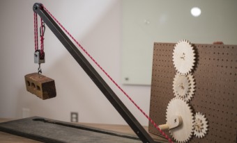 Three differently sized gears are affixed to a wooden background with holes in it.  The gears are connect to a long pulley that is supporting a wooden block. 