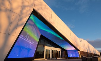 Photo of the Canada Science and Technology Museum's façade.  The building is a beige triangular shape with a series of screens showing differently colored lights over the main entrance doors. 