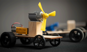 Gros plan sur un petit véhicule en bois avec des roues noires et une hélice jaune fixée à un moteur gris.