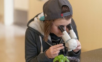 A child wearing a backwards cap and a grey fleece jacket is bent over a microscope looking at a soil sample. The room behind the child is out of focus.