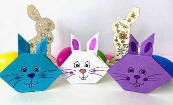 Three origami paper rabbit faces are lined up, sitting on a white background. One is blue, one is white, and one is purple; each has a cartoon-style face in black ink. Behind the paper rabbits are a collection of brightly coloured eggs and decorative wooden rabbits.
