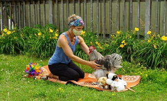 A woman wearing a face mask kneels on a yoga mat on the grass. She is reaching for a grey rooster who stands next to her on the mat; a series of toys are arranged around them. 