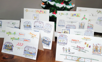 Eight brightly-coloured holiday cards are arranged on a wooden tabletop; each card features different aviation-themed images. Christmas décor is visible in the foreground and background of the image.