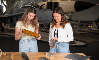 Two girls stand in front of a wooden table covered with materials used to fashion airplanes.  The girl on the left has long blonde hair, a pale tshirt and jeans. She is holding a rectangular prism. The girl on the right has long brown hair and is holding a large grey cylinder.  In the background are the noses of two Canadian jets in the aviation museum collection. 