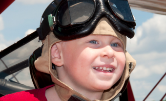 A smiling child wears a red shirt, a tan aviator helmet and large black aviator goggles. Behind him there is a cloudy blue sky and the top of a red biplane wing.