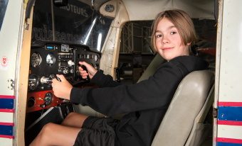 A blond boy with chin length hair and a black long sleeved top and black shorts sits at the controls of a Cesna aircraft.