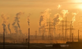 Telephone poles and electric towers in the foreground and a hazy skyline with multiple stacks releasing pollution into a golden sky in the background.