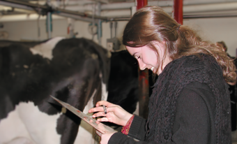 A teenaged girl smiles as she looks down at a clipboard. Cows are visible in the background.
