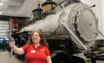 Une employée de musée se tient devant une grande locomotive à vapeur grise et noire. Elle porte un écouteur et sourit à l'écran d'un téléphone intelligent qu'elle tient par un cardan dans son bras tendu.