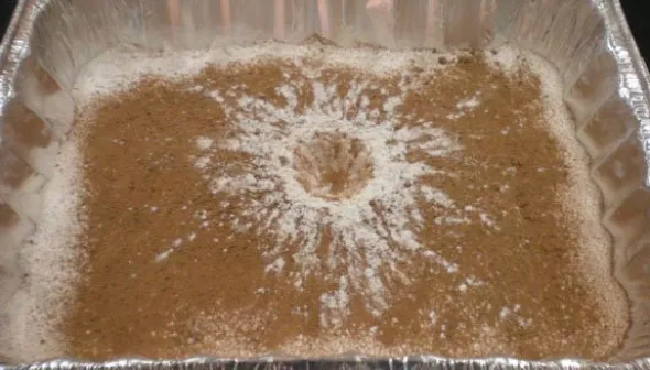 Un cratère formé de cacao et de farine, dans un plat rectangulaire jetable. Le cratère est brun et blanc, avec des traînées blanches laissées par la farine qui a jailli du centre. 
