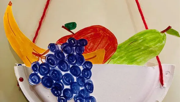 Un bricolage d’un panier de fruits fait à partir d’une assiette en papier. Le panier bricolé est enfilé et suspendu avec une corde rouge. Des images faites à la main de fruits colorés ressortent du dessus du panier.