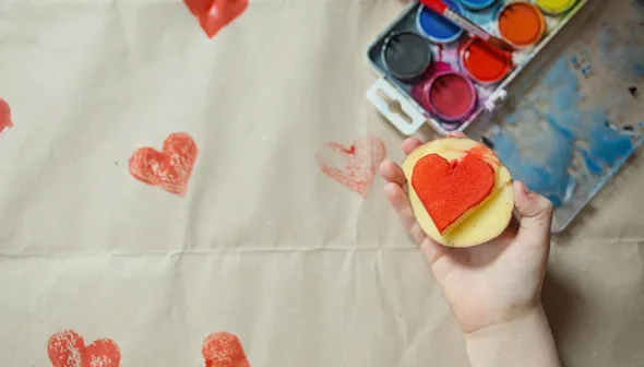 Une main d’enfant tient une estampe en patate avec un cœur peint en rouge. En arrière-plan, un plateau de peinture et plusieurs cœurs rouges estampés sont visibles contre le fond blanc.