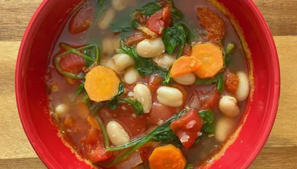 Un bol de soupe rouge est posé sur une planche à découper en bois. Le bol est rempli de soupe dans laquelle on voit des épinards, des tomates, des haricots blancs et des carottes.