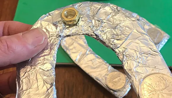 A hand holds a homemade horseshoe, preparing to toss it on to a tabletop horseshoe game. The horseshoes are covered in tinfoil and weighed with metal objects such as coins and washers.