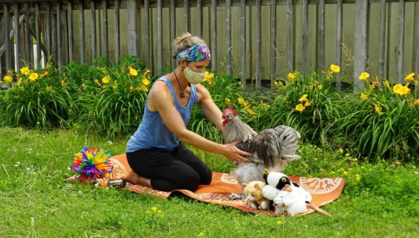 Une femme portant un masque facial, est agenouillée sur un tapis de yoga posé sur l'herbe. Elle tend la main vers un coq gris qui se trouve à côté d'elle sur le tapis. Une série de jouets sont disposés autour d’eux.