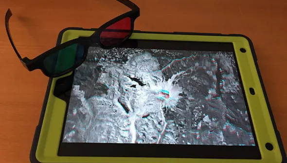 A pair of 3D glasses, with one red lens and one blue lens, leaning on an iPad in a green case.  The iPad has an anaglyph satellite image of Mount St Helens, for viewing with the glasses, on its screen.