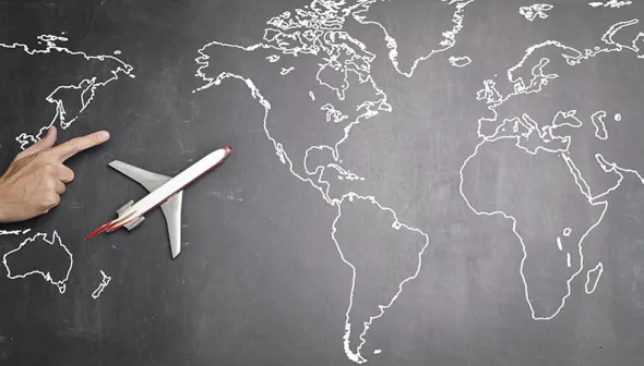 Modèle d'un avion de ligne, positionné sur un tableau noir sur lequel une carte du monde est dessinée à la craie blanche. À côté de l'avion se trouve une main avec un doigt qui indique la direction de vol de l'avion. 